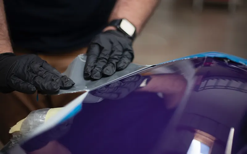 A paint booth technician is applying chemical on the surface of a purple car